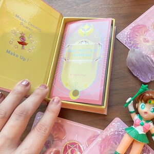 Puede incluir: Una caja rosa y amarilla que contiene una gu&iacute;a Moon Hero Tarot con el texto Moon Cosmic Power, Make Up! impreso en el interior. El set incluye cartas de tarot rosas con motivos de estrellas y una peque&ntilde;a figura con un traje verde y blanco.