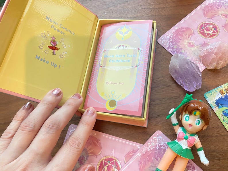 Puede incluir: Una caja rosa y amarilla que contiene una gu&iacute;a Moon Hero Tarot con el texto Moon Cosmic Power, Make Up! impreso en el interior. El set incluye cartas de tarot rosas con motivos de estrellas y una peque&ntilde;a figura con un traje verde y blanco.