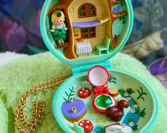 May include: A mint green plastic locket playset that opens to reveal a miniature treehouse interior with a small doll, a table, and a chair. The bottom section features a green gem, a brown squirrel figure, red mushrooms, and a gold chain.