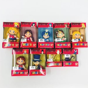 Op de afbeelding: Een verzameling Sailor Moon-personagefiguren in hun originele verpakking. Elke figuur zit in een doorzichtige plastic hoes met een rode kartonnen doos. De figuren hebben levendige kleuren en gedetailleerde ontwerpen.