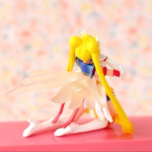 May include: A Sailor Moon figurine kneeling in a pose with long blonde hair styled in twin buns and long pigtails. The character wears a white and blue outfit with red accents and features translucent, wing-like appendages extending from the back.