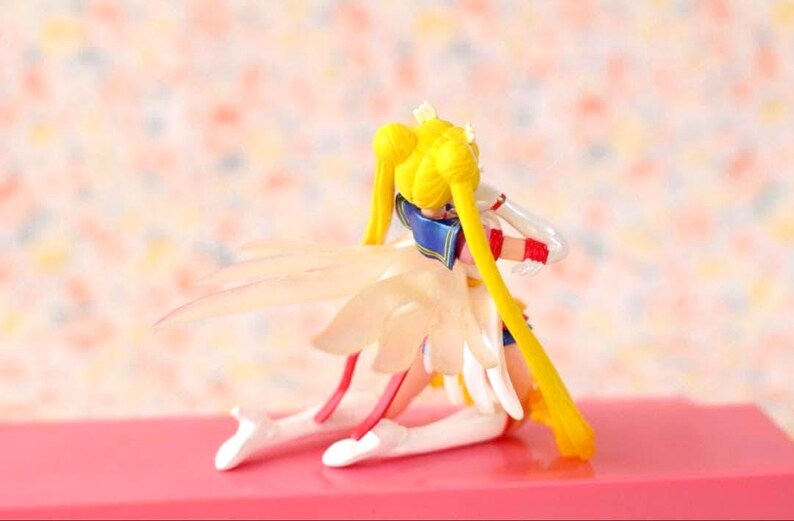 May include: A Sailor Moon figurine kneeling in a pose with long blonde hair styled in twin buns and long pigtails. The character wears a white and blue outfit with red accents and features translucent, wing-like appendages extending from the back.