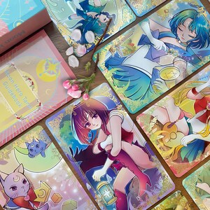 Puede incluir: Una baraja de tarot Moon Hero que incluye una gu&iacute;a rosa con el texto Moon Hero Tarot Guidebook by Sharon Tsui, junto a varias cartas ilustradas con personajes de estilo anime, bordes dorados de filigrana y un gato.