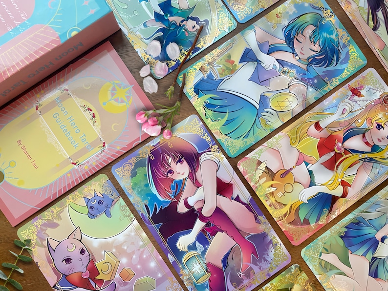 Puede incluir: Una baraja de tarot Moon Hero que incluye una gu&iacute;a rosa con el texto Moon Hero Tarot Guidebook by Sharon Tsui, junto a varias cartas ilustradas con personajes de estilo anime, bordes dorados de filigrana y un gato.