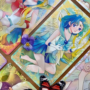 Puede incluir: Una carta de tarot rectangular que muestra un personaje de estilo anime con cabello azul, vistiendo un atuendo de marinero blanco y azul con un emblema de coraz&oacute;n, enmarcado por un borde dorado ornamentado, con el n&uacute;mero romano XIV visible en la parte inferior.