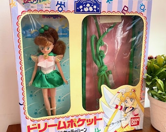 May include: A vintage Sailor Moon doll in its original box. The doll has brown hair, a green skirt, and a pink top. The box is decorated with Japanese text and illustrations of Sailor Moon characters. The box also contains a green rope.
