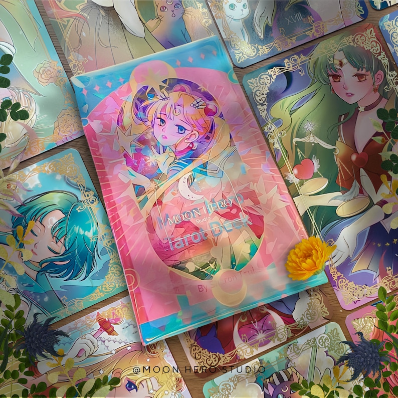 Puede incluir: Una caja de cartas de tarot con una ilustraci&oacute;n central de un personaje rubio con dos mo&ntilde;os, rodeado de acentos hologr&aacute;ficos rosas y azules. El texto dice Moon Hero Studio Tarot Deck. Las cartas circundantes muestran varios personajes.