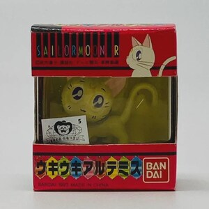 Op de afbeelding: Een Sailor Moon R figuur van een gele kat genaamd Artemis, in een rode en transparante plastic doos. De doos heeft Japanse tekst en een witte kat illustratie. Het figuur is een verzamelobject van Bandai.