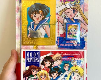 May include: A Sailor Moon S Moon Princess Bromideset featuring a gold-bordered card with a character illustration, a small blue magnet, and a white collection card case. The packaging displays various anime characters and Japanese text throughout.