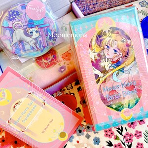 Puede incluir: Un mazo de tarot Moon Hero y su gu&iacute;a con ilustraciones estilo anime en tonos rosa y azul. Incluye un bot&oacute;n circular con un gato y el texto Mooniemons, adem&aacute;s de una peque&ntilde;a tarjeta azul con forma de coraz&oacute;n.