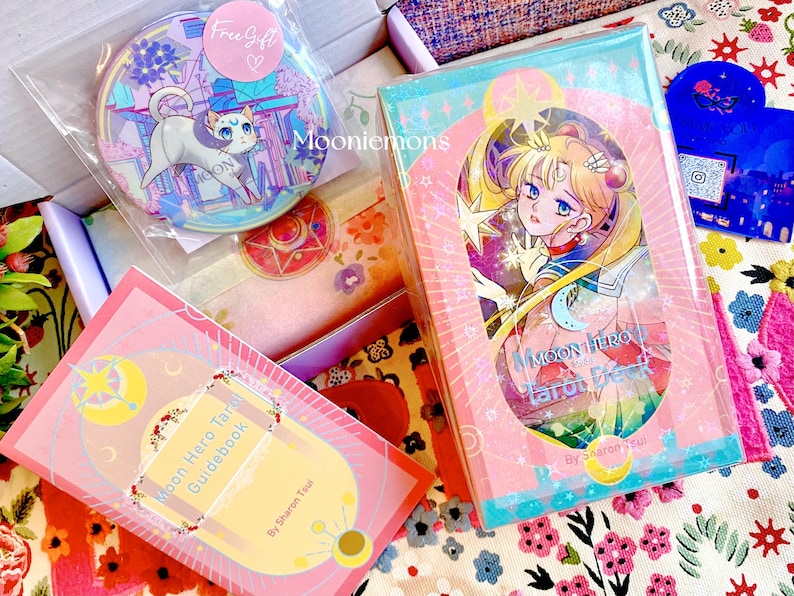 Puede incluir: Un mazo de tarot Moon Hero y su gu&iacute;a con ilustraciones estilo anime en tonos rosa y azul. Incluye un bot&oacute;n circular con un gato y el texto Mooniemons, adem&aacute;s de una peque&ntilde;a tarjeta azul con forma de coraz&oacute;n.