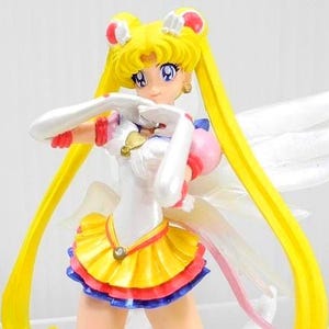 May include: A Sailor Moon anime character figurine featuring long yellow hair in twin tails, wearing a white leotard with a yellow pleated skirt, red and blue trim, white gloves, and translucent wings. The figure has large purple eyes and a gold heart emblem.