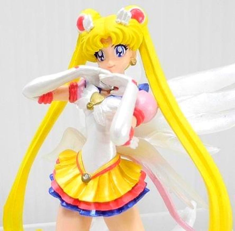 May include: A Sailor Moon anime character figurine featuring long yellow hair in twin tails, wearing a white leotard with a yellow pleated skirt, red and blue trim, white gloves, and translucent wings. The figure has large purple eyes and a gold heart emblem.