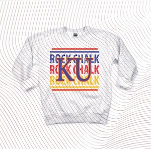 May include: A light gray sweatshirt with long sleeves. The front features the text "ROCK CHALK" in blue, red, and yellow, with a large "KU" in blue and yellow. The design is centered on the chest.