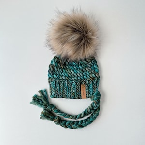 Könnte beinhalten: Tealblauer Strickmütze mit einem braunen Kunstfellbommel und langen geflochtenen Bändern in Tealblau. Die Mütze hat ein braunes Leder-Etikett mit dem Text "GO NORTH" darauf.