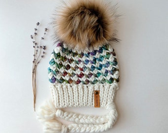 Puede incluir: Gorro de punto blanco con un patrón de rombos de colores, un pompón de piel sintética marrón y una etiqueta de cuero marrón que dice "Go North". El gorro tiene largas cintas trenzadas blancas.