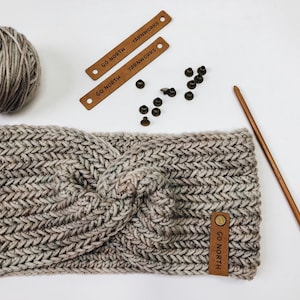 May include: A hand-knitted headband in shades of gray and brown, with a twisted design. The headband is accompanied by a ball of yarn, leather tags with "GO NORTH YARWORKS" and metal rivets. A wooden crochet hook is also present.
