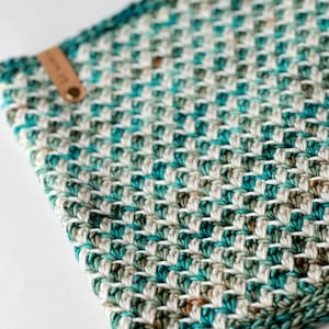 Puede incluir: Un cuadrado de crochet de color turquesa, blanco y marrón con una etiqueta de cuero adjunta. El cuadrado de crochet está hecho con un patrón de chevron.