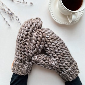 K&ouml;nnte beinhalten: Ein Paar braune und wei&szlig;e Strickhandschuhe mit einem Lederriemen. Die Handschuhe werden von einer Person gehalten, die ein schwarzes Langarmshirt tr&auml;gt.