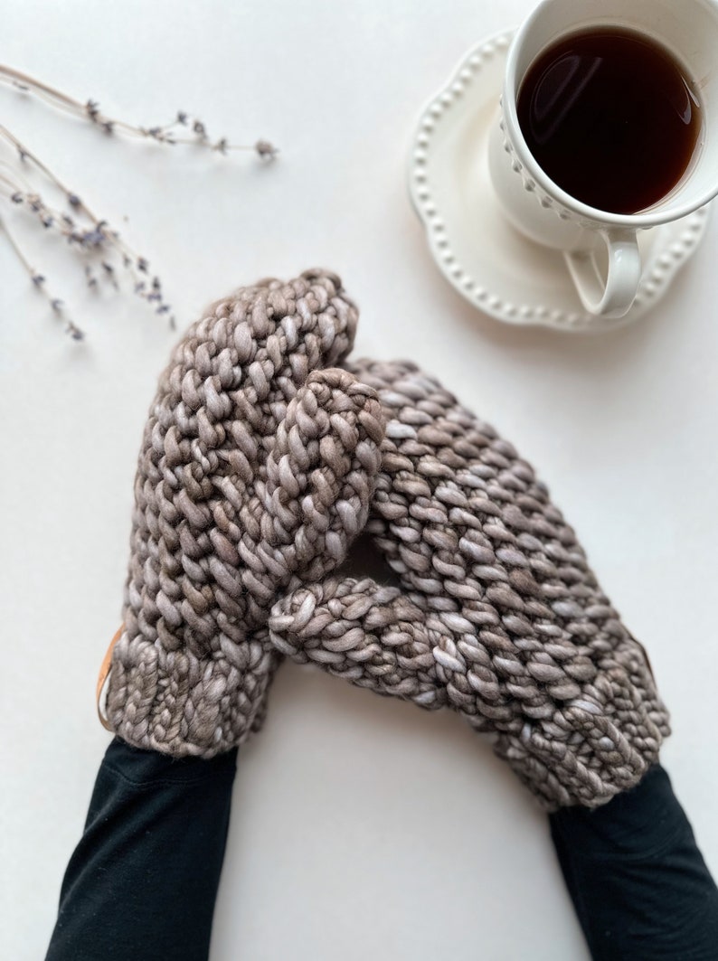 K&ouml;nnte beinhalten: Ein Paar braune und wei&szlig;e Strickhandschuhe mit einem Lederriemen. Die Handschuhe werden von einer Person gehalten, die ein schwarzes Langarmshirt tr&auml;gt.