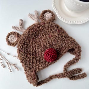 Puede incluir: Un gorro de ganchillo marrón con características de reno, incluyendo cuernos, orejas y una nariz roja. El gorro tiene una apariencia texturizada y orejeras. Probablemente para un bebé o niño.