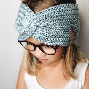 Puede incluir: Una diadema de punto azul claro con un diseño de nudo retorcido. La diadema tiene un aspecto texturizado y una pequeña etiqueta de cuero marrón con el texto "GO NORTH". La diadema es usada por una persona que lleva gafas.