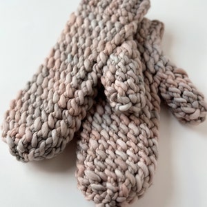K&ouml;nnte beinhalten: Ein Paar gestrickte F&auml;ustlinge in einem hellbraunen und rosafarbenen Farbschema. Die F&auml;ustlinge sind aus einem dicken Garn gefertigt und haben ein strukturiertes Stichmuster.