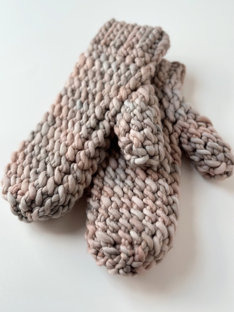 K&ouml;nnte beinhalten: Ein Paar gestrickte F&auml;ustlinge in einem hellbraunen und rosafarbenen Farbschema. Die F&auml;ustlinge sind aus einem dicken Garn gefertigt und haben ein strukturiertes Stichmuster.