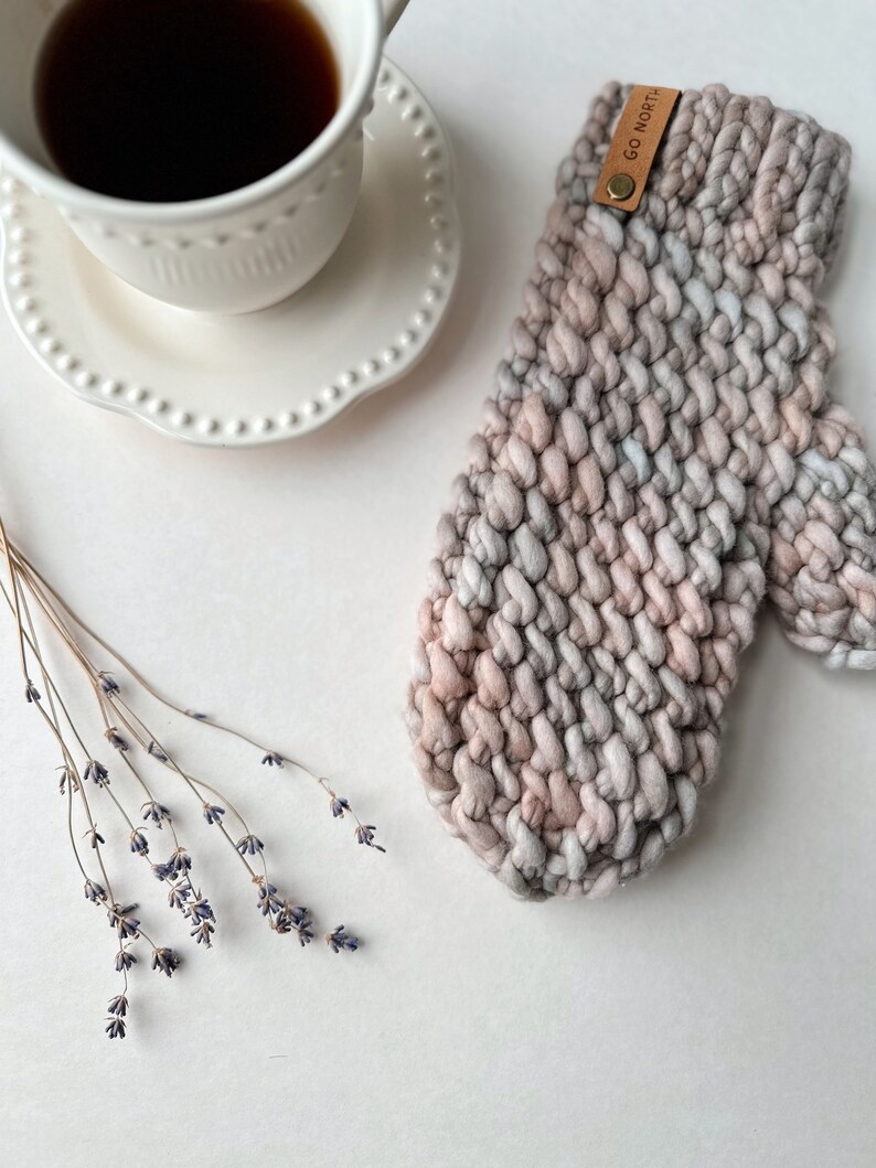 K&ouml;nnte beinhalten: Eine dicke Strickm&uuml;tze in Rosa-, Grau- und Wei&szlig;t&ouml;nen mit einem braunen Lederanh&auml;nger mit der Aufschrift "GO NORTH".