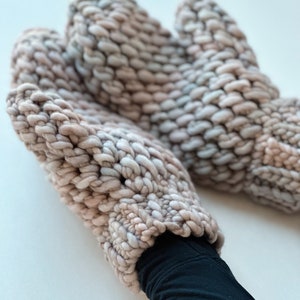 K&ouml;nnte beinhalten: Ein Paar dicke Strickhandschuhe in einem hellrosa und grauen Farbschema. Die Handschuhe sind aus dickem Garn gefertigt und haben ein strukturiertes, Zopfmuster.