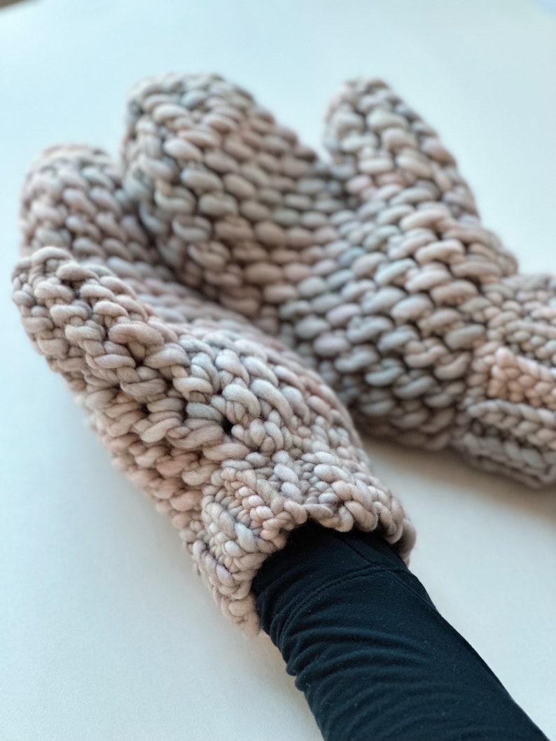 K&ouml;nnte beinhalten: Ein Paar dicke Strickhandschuhe in einem hellrosa und grauen Farbschema. Die Handschuhe sind aus dickem Garn gefertigt und haben ein strukturiertes, Zopfmuster.