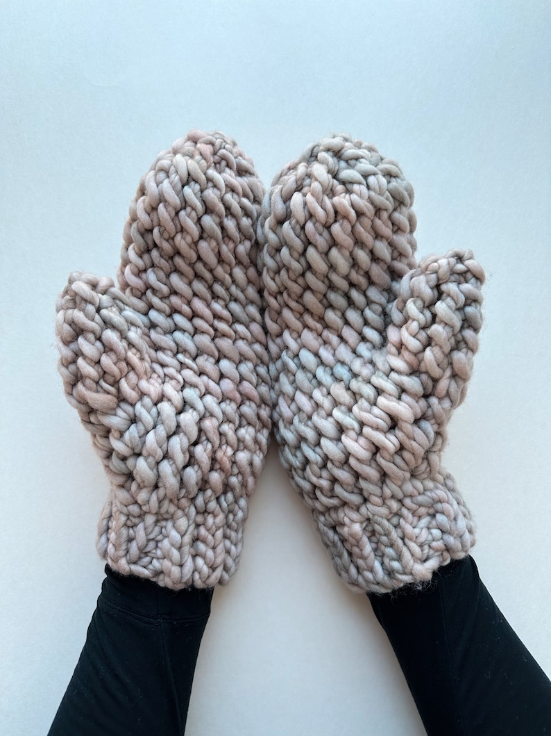 K&ouml;nnte beinhalten: Ein Paar dicke Strickhandschuhe in einem hellrosa und grauen Farbschema. Die Handschuhe sind aus dickem Garn gefertigt und haben eine strukturierte, volumin&ouml;se Optik.