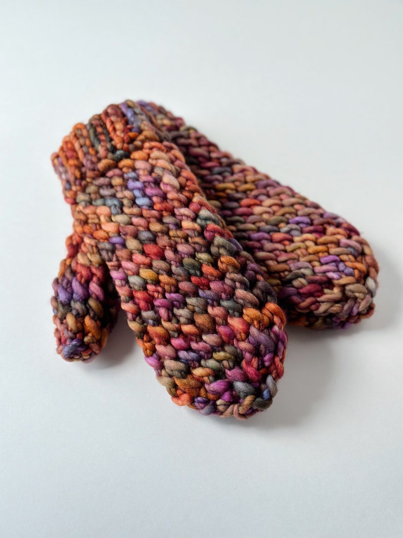 K&ouml;nnte beinhalten: Ein Paar bunte Strickhandschuhe mit einem strukturierten Muster. Die Handschuhe sind aus verschiedenen Braunt&ouml;nen, Lila und Orange gefertigt.