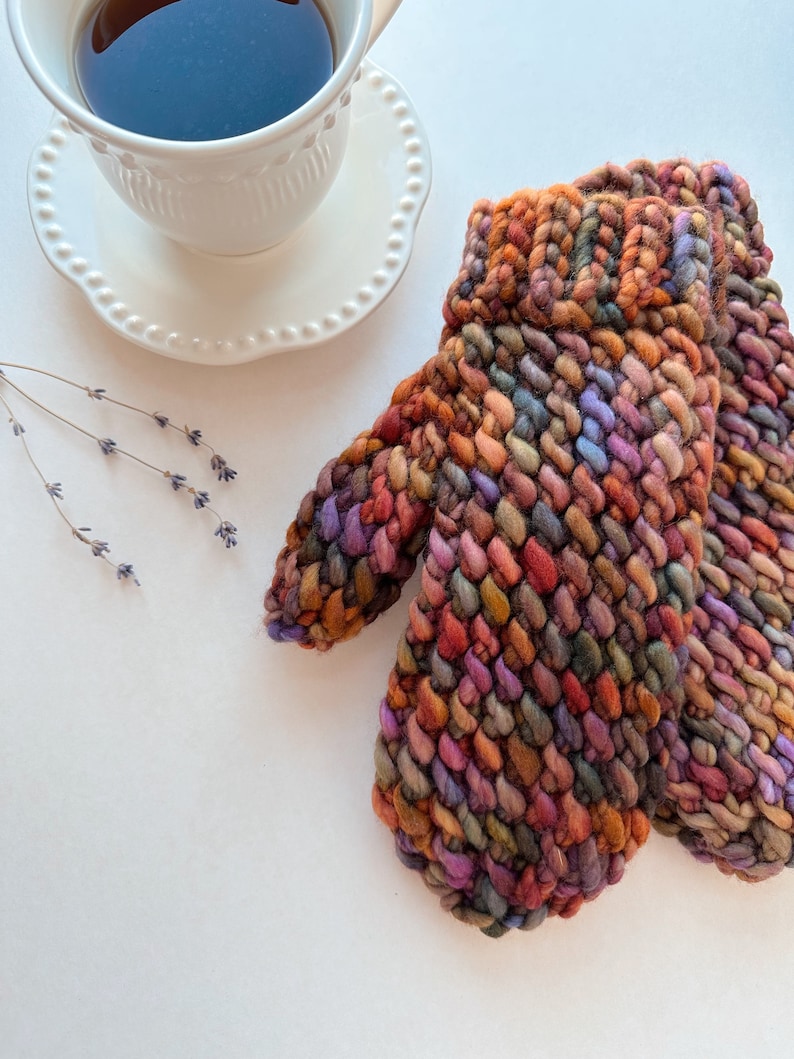 K&ouml;nnte beinhalten: Ein Paar bunte Strickhandschuhe mit einem strukturierten Muster. Die Handschuhe sind aus verschiedenen Braunt&ouml;nen, Orange, Lila und Gr&uuml;n gefertigt.