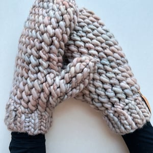 K&ouml;nnte beinhalten: Ein Paar dicke Strickhandschuhe in einem hellrosa und grauen Farbschema. Die Handschuhe sind aus dickem Garn gefertigt und haben eine strukturierte Optik.