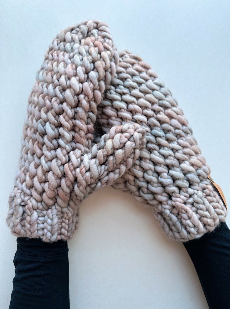 K&ouml;nnte beinhalten: Ein Paar dicke Strickhandschuhe in einem hellrosa und grauen Farbschema. Die Handschuhe sind aus dickem Garn gefertigt und haben eine strukturierte Optik.