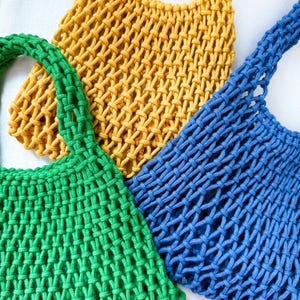 May include: Three handmade macrame tote bags in vibrant colors: green, yellow, and blue. The bags feature a woven net design with sturdy handles. The bags are ideal for carrying groceries or other items.