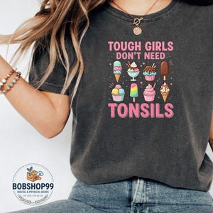 May include: A charcoal grey short-sleeve t-shirt featuring the pink text "TOUGH GIRLS DON'T NEED TONSILS" above and below a graphic illustration of eight colourful ice cream treats, including cones, sundaes, and popsicles in various pastel shades.