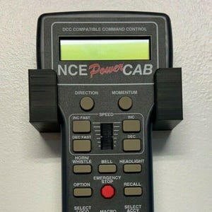 May include: Black NCE Power Cab DCC compatible command control unit with numerous buttons and a green display screen. Features buttons for direction, speed, and emergency stop. Includes a black mounting bracket.
