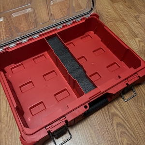 May include: A red plastic tool box with a clear lid. The interior has compartments and a black foam divider. The box has a black handle and metal latches. The brand name "Milwaukee" is visible on the lid.