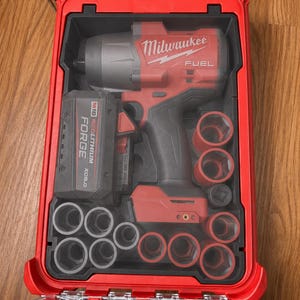 May include: A red Milwaukee Fuel impact wrench with an M18 Redlithium Forge XC6.0 battery, housed in a red plastic carrying case with a clear lid. The set includes multiple black impact sockets arranged in custom-molded slots within the case.