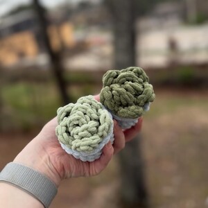 May include: Two handmade, crocheted items in the image. One is light green, the other is olive green. Both are round with a textured, looped design. The base of each item is a light blue-gray color. The items are held in a hand.