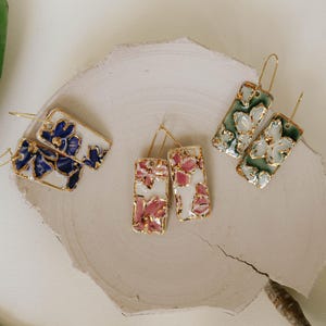 May include: Four pairs of rectangular dangle earrings with floral designs. The earrings feature gold-colored frames and are decorated with blue, pink, and green floral patterns on a white background. The earrings are displayed on a light-colored, textured surface.