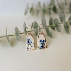 Puede incluir: Pendientes de cerámica con base blanca, diseño floral azul y detalles dorados. Los pendientes tienen forma rectangular con la parte superior redondeada y ganchos dorados. El diseño incluye un jarrón con flores.