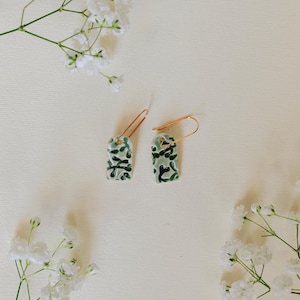 Puede incluir: Dos pendientes rectangulares con base verde claro y un patrón abstracto verde oscuro. Los pendientes tienen ganchos para las orejas de color cobre. Se exhiben sobre una superficie color crema, rodeados de flores blancas.