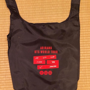May include: Black tote bag with red text that reads "ARIRANG BTS WORLD TOUR." The bag has two handles and a rectangular design. The text includes the names of the band members and a checkmark.