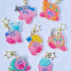 以下が含まれることがあります： 様々なコスチュームデザインのピンク色のカービィキャラクターのアクリルキーホルダーのコレクション。各キーホルダーには、金色の星型の留め金が付いています。デザインには、忍者、火、その他のテーマの衣装が含まれており、鮮やかな色と詳細なアートワークが特徴です。