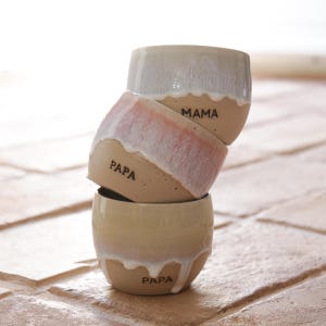 Könnte beinhalten: Ein Stapel von drei Keramikbechern mit beigem Boden und weißer, tropfender Glasur. Der obere Becher ist weiß, der mittlere Becher ist rosa und der untere Becher ist beige. Das Wort "MAMA" steht auf dem mittleren Becher und "PAPA" auf den anderen beiden.