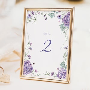 May include: A rectangular gold frame with a floral design and the number 2. The frame is on a white surface. The floral design includes purple flowers and green leaves. The text "Table No." is above the number 2.