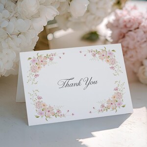 Puede incluir: Una tarjeta blanca plegada con las palabras "Thank You" en escritura elegante, enmarcada por un delicado borde floral en tonos rosa y melocotón. La tarjeta está sobre una superficie blanca, con un fondo de flores blancas y rosas.